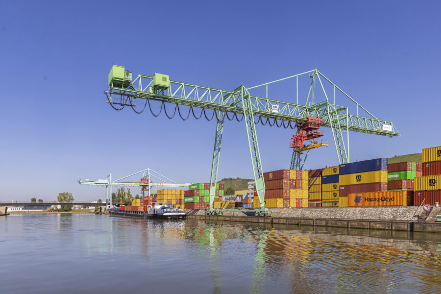 Großer Hafenkran über gestapelten Containern am Wasser, blauer Himmel.