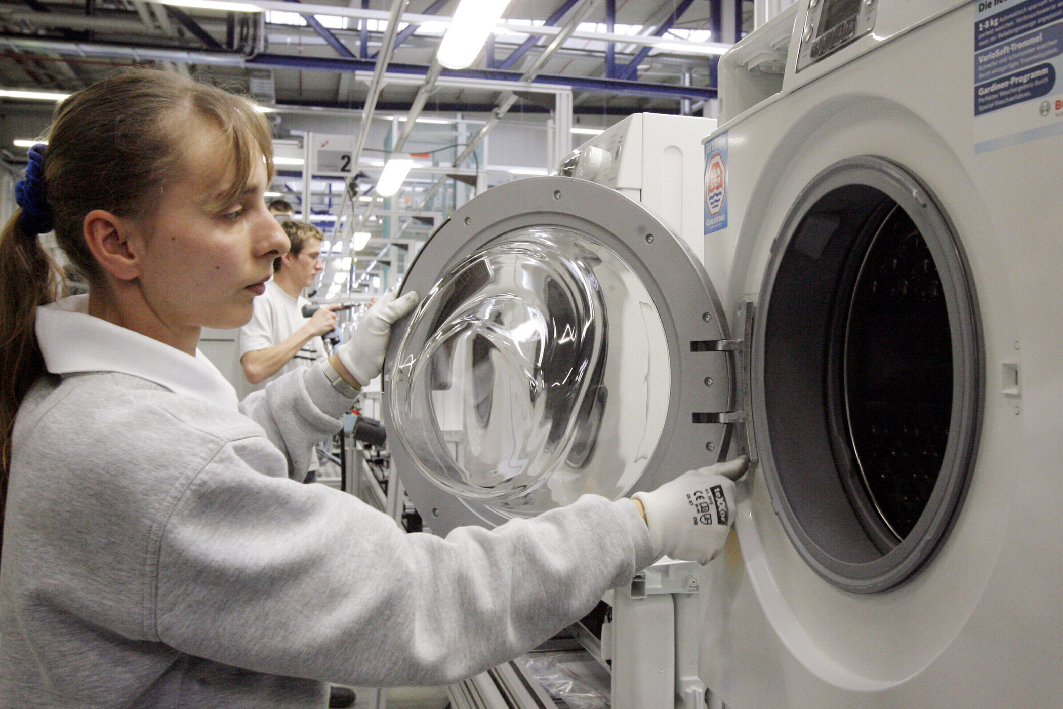 Zwei Personen arbeiten an einer Waschmaschine in einer Fabrikhalle.