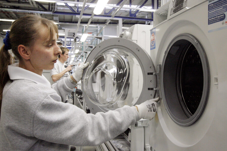 Zwei Personen arbeiten an einer Waschmaschine in einer Fabrikhalle.