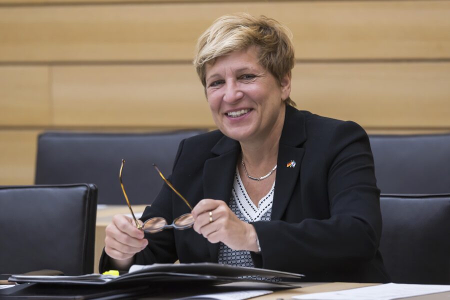 Lächelnde Person mit Brille in der Hand, sitzt an einem Tisch mit Unterlagen.
