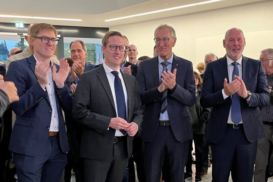 Vier Männer in Anzügen klatschen in einem Raum mit Publikum.