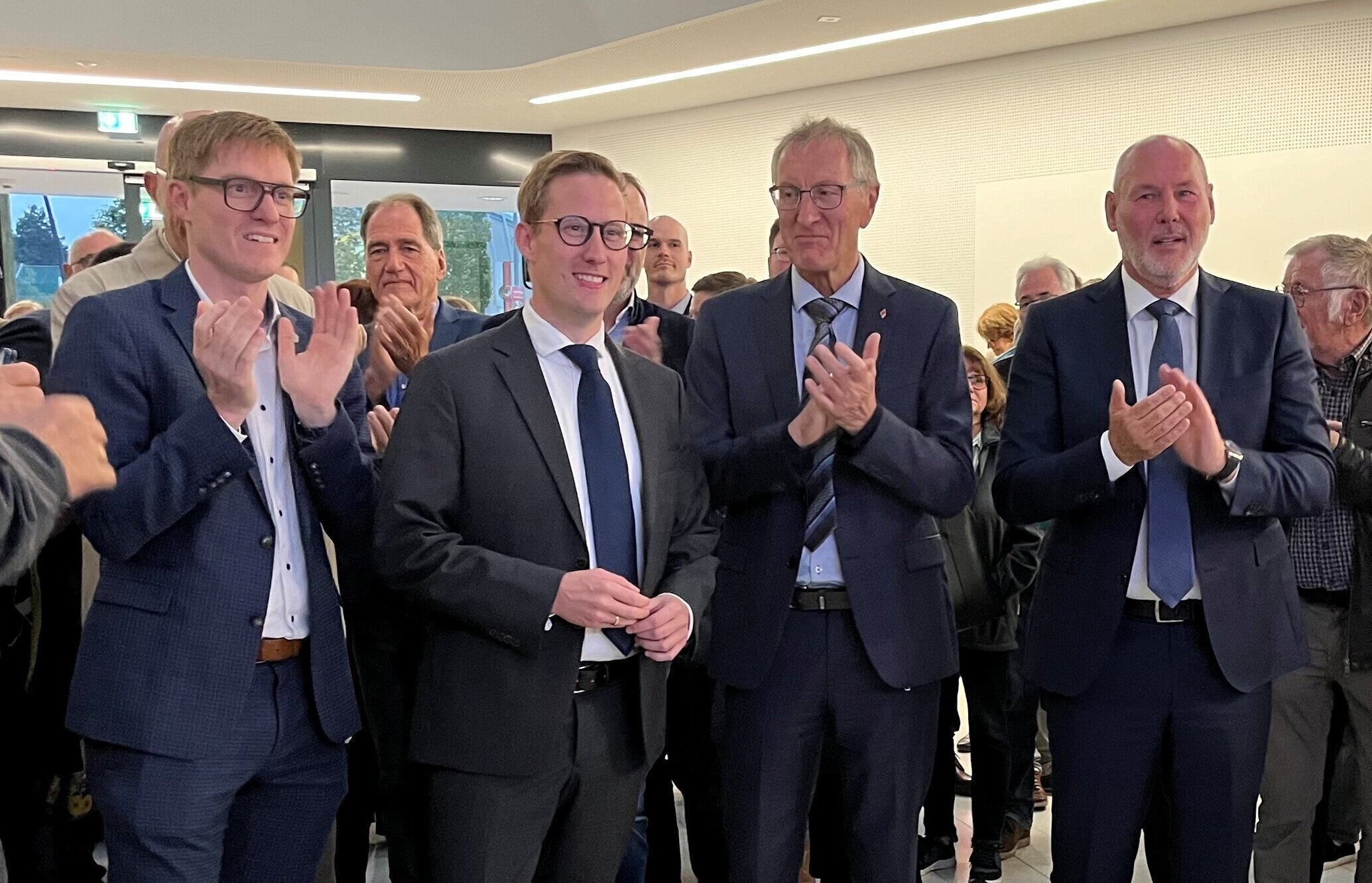 Vier Männer in Anzügen klatschen in einem Raum mit Publikum.