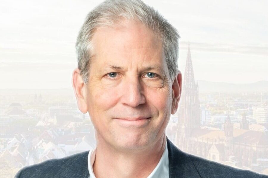 Mann mit grauem Haar lächelt, im Hintergrund eine Stadtansicht mit Kirchturm.