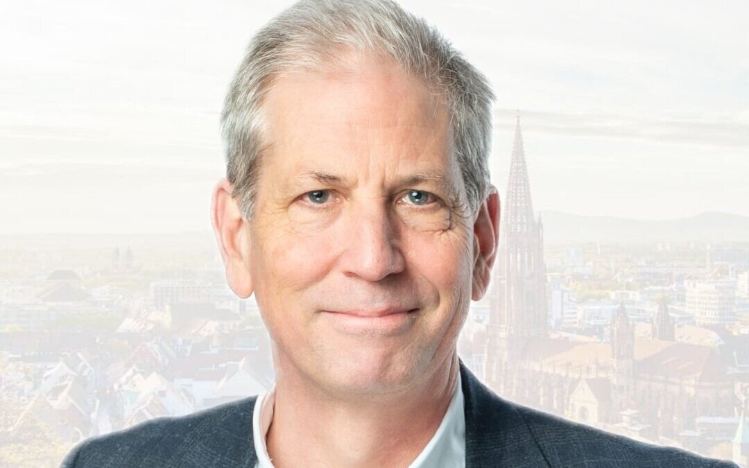 Mann mit grauem Haar lächelt, im Hintergrund eine Stadtansicht mit Kirchturm.