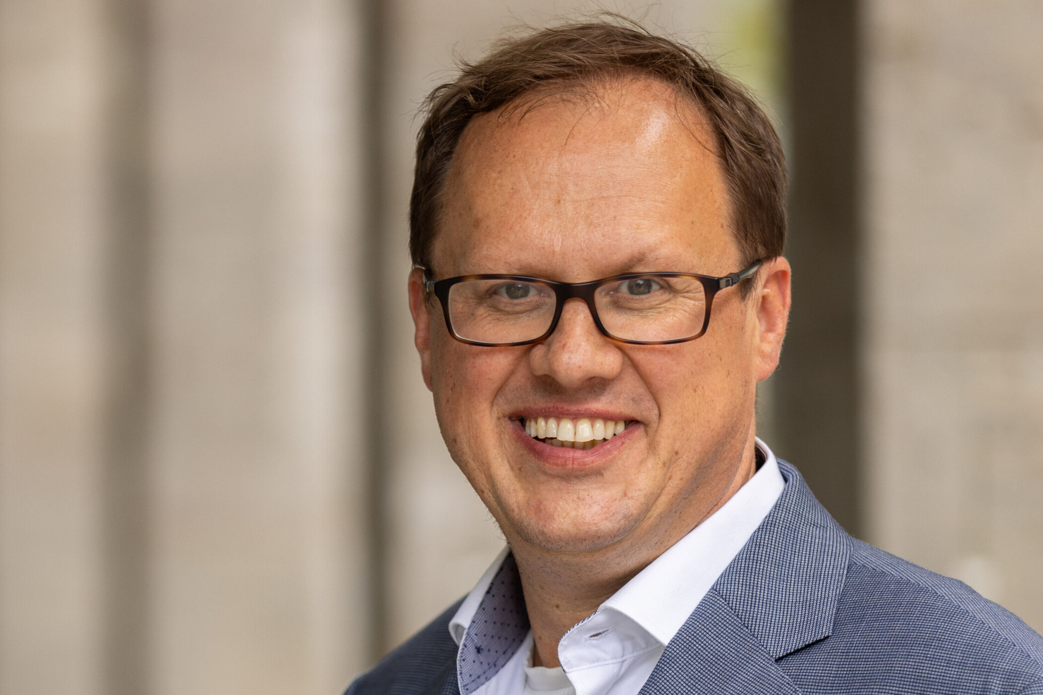 Lächelnder Mann mit Brille und blauem Sakko vor unscharfem Hintergrund.
