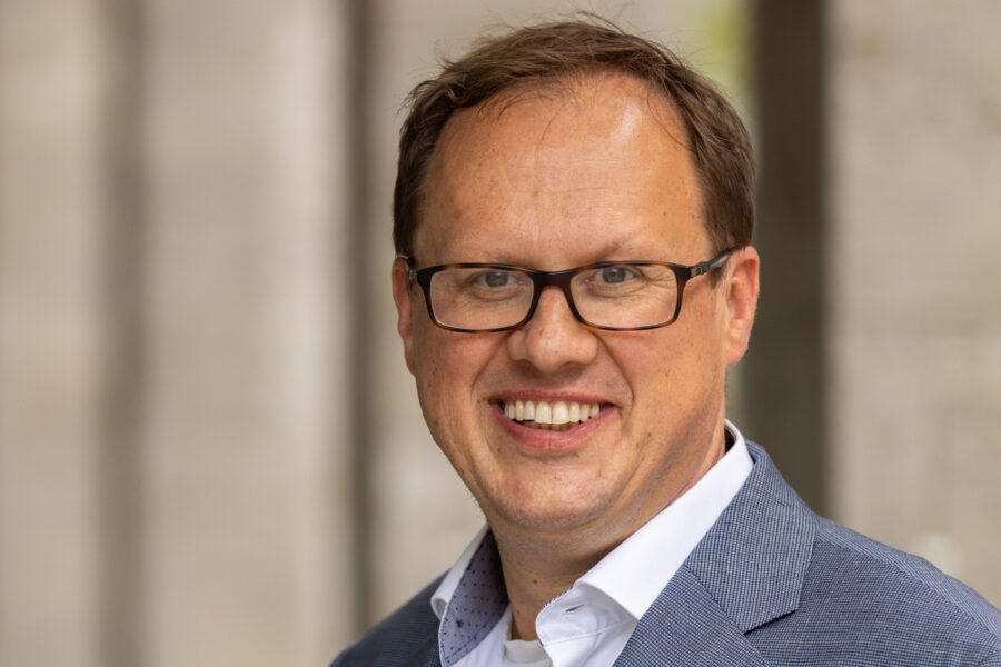 Lächelnder Mann mit Brille und blauem Sakko vor unscharfem Hintergrund.
