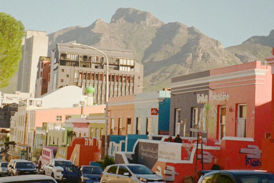 Bunte Häuserreihe vor Bergen, Autos und Menschen auf der Straße.