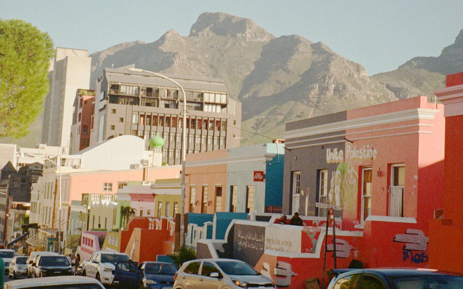 Bunte Häuserreihe vor Bergen, Autos und Menschen auf der Straße.