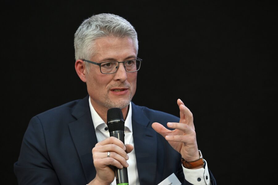 Mann mit Brille und Bart spricht in ein Mikrofon, trägt Anzug und Hemd.