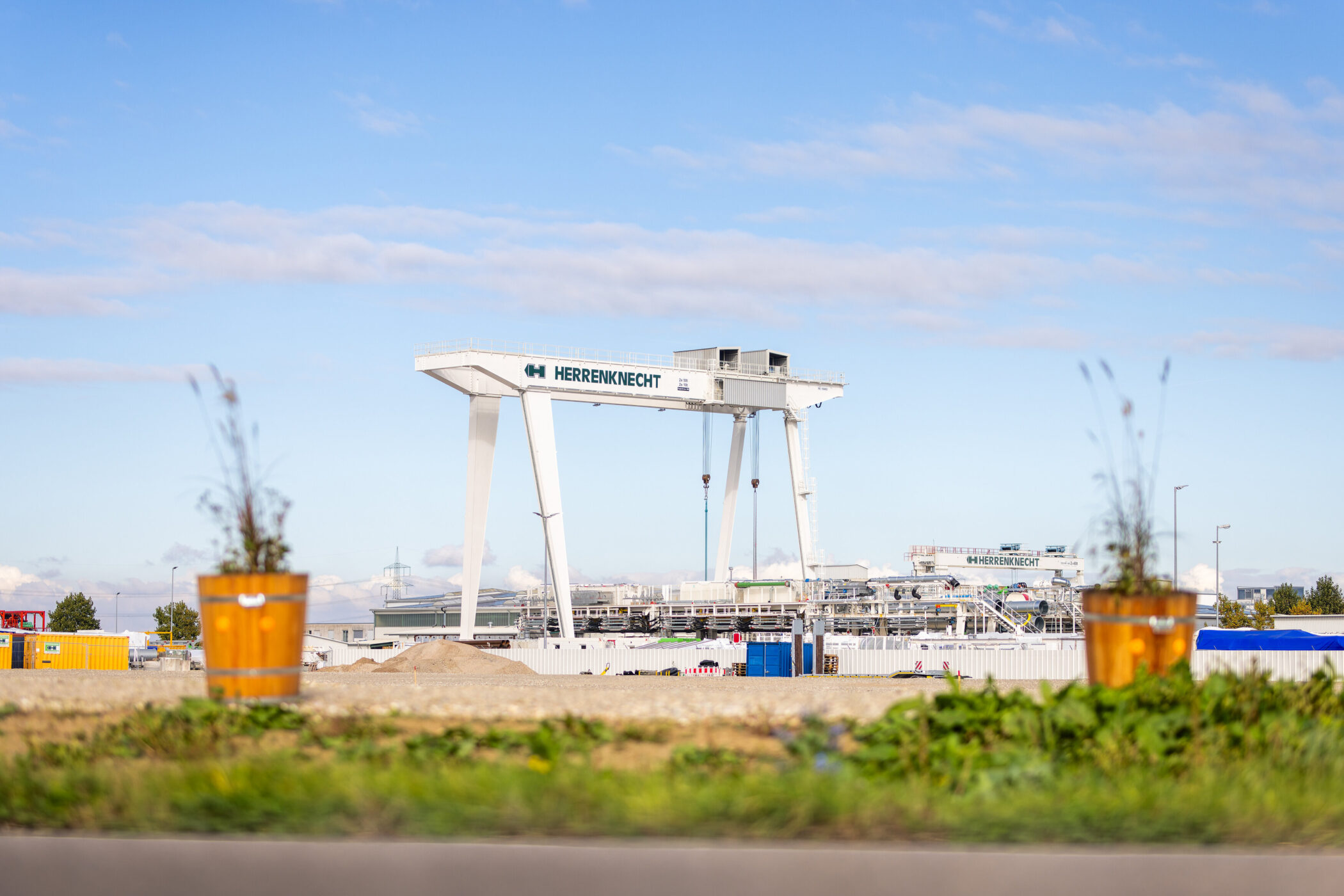 Großer Kran mit "HERRENKNECHT"-Schriftzug auf Baustelle, blauer Himmel.