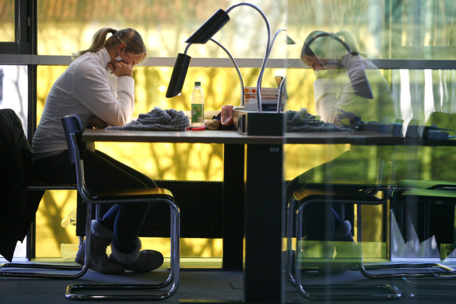 Person sitzt an einem Tisch, liest, mit Lampe und Getränkeflasche. Fenster im Hintergrund.