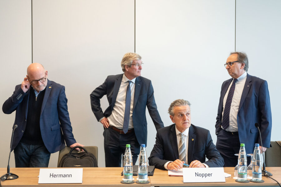 Vier Männer in Anzügen an einem Tisch mit Namensschildern und Wasserflaschen.