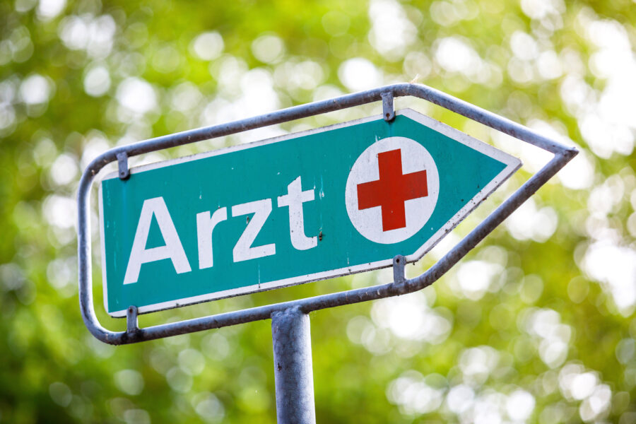 Grünes Schild mit rotem Kreuz und Text "Arzt".