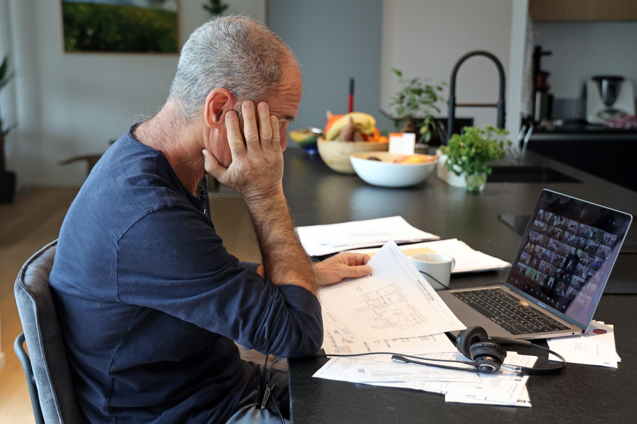 Mann sitzt am Tisch, betrachtet Dokumente, Laptop mit Videokonferenz, Kopfhörer daneben.