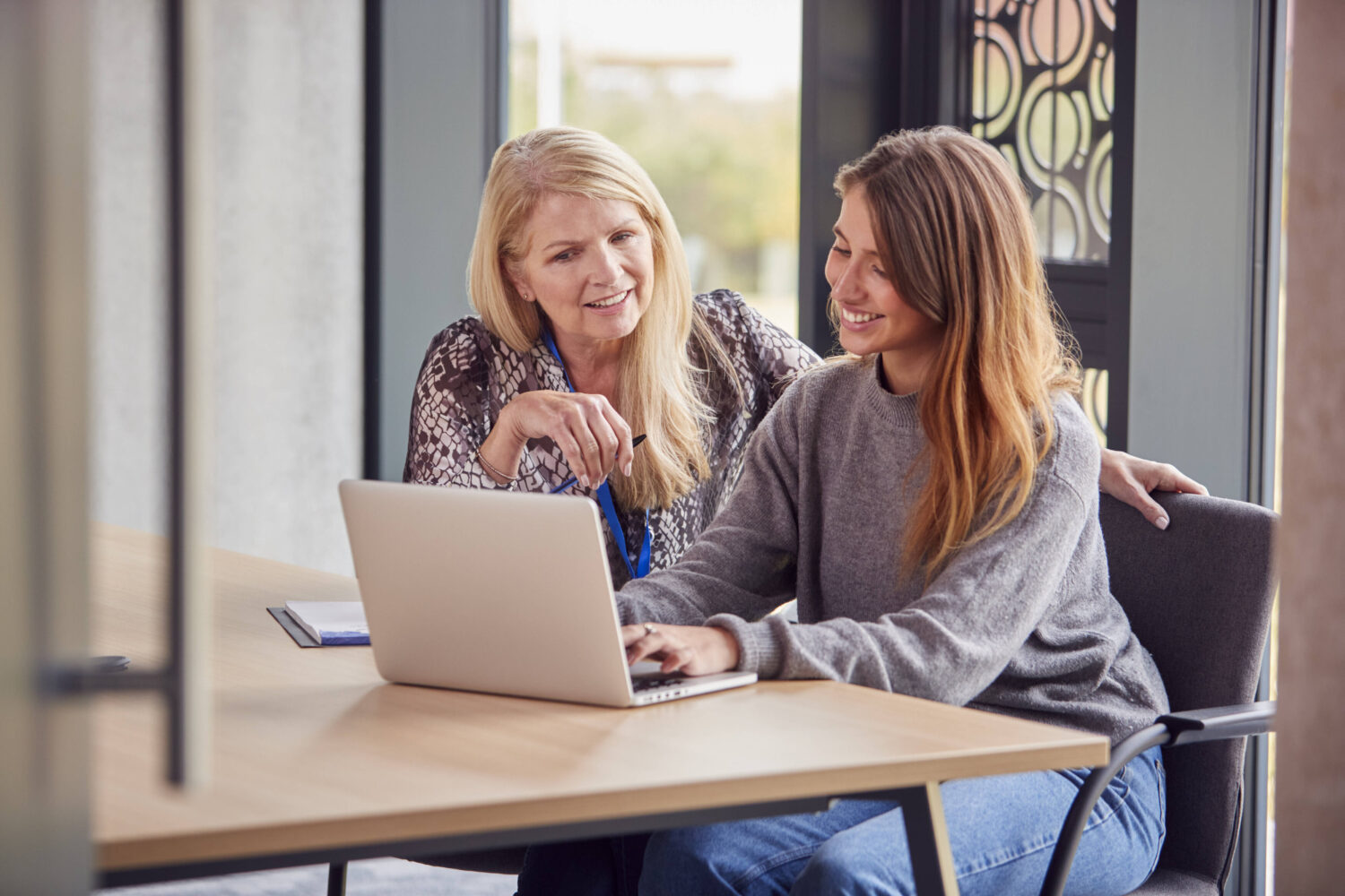 Zwei Frauen sitzen an einem Tisch mit Laptop, lächelnd und in Gespräch vertieft.