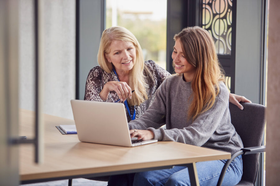 Zwei Frauen sitzen an einem Tisch mit Laptop, lächelnd und in Gespräch vertieft.