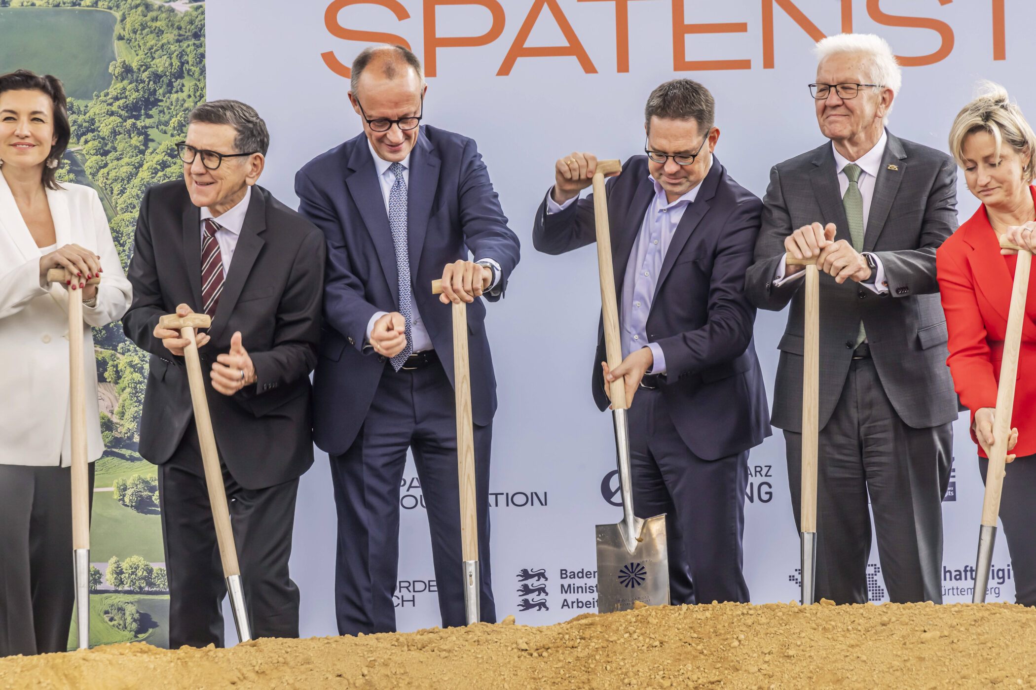 Sechs Personen in Anzügen halten Schaufeln bei einer Zeremonie.