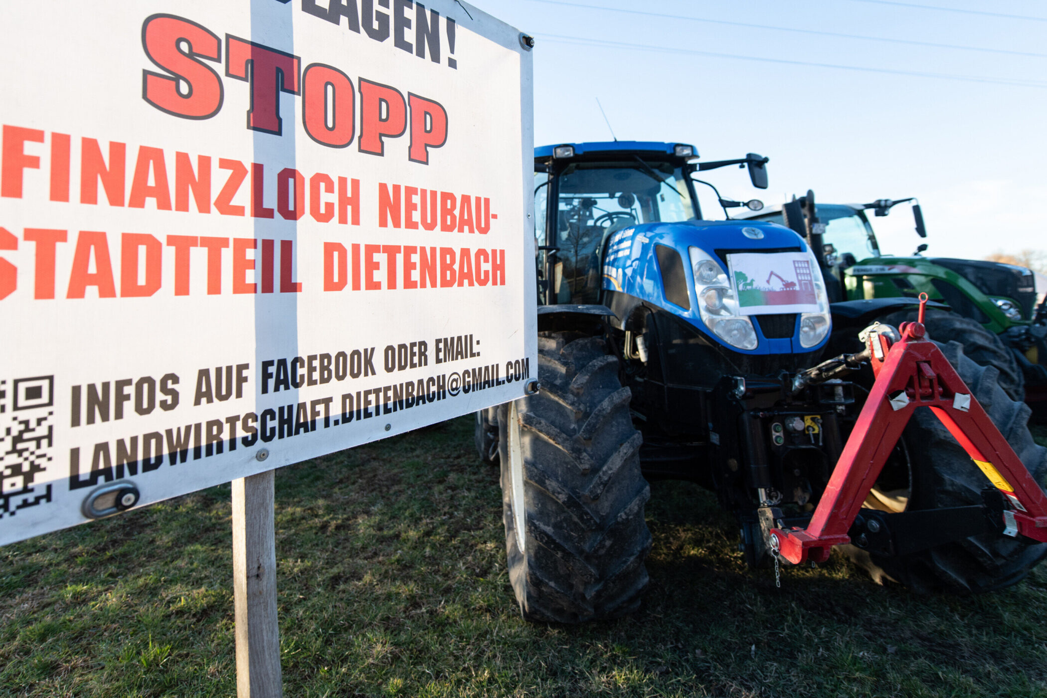 Schild mit Protesttext gegen Neubau, zwei Traktoren im Hintergrund.