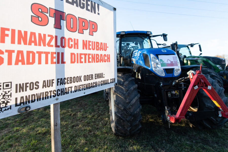 Schild mit Protesttext gegen Neubau, zwei Traktoren im Hintergrund.