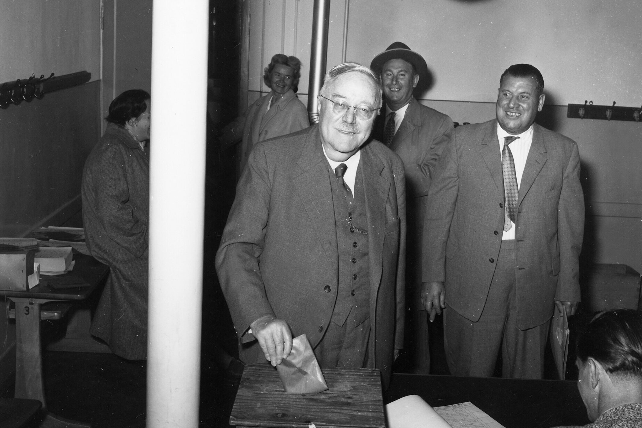 Sechs Männer in Anzügen, einer wirft einen Umschlag in eine Wahlurne.