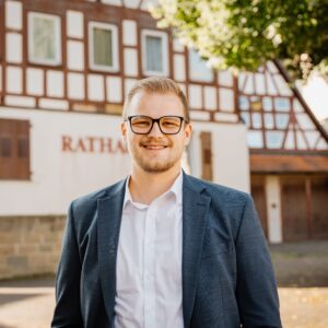 Mann mit Brille und Anzug vor Fachwerkhaus mit "Rathaus"-Schriftzug.