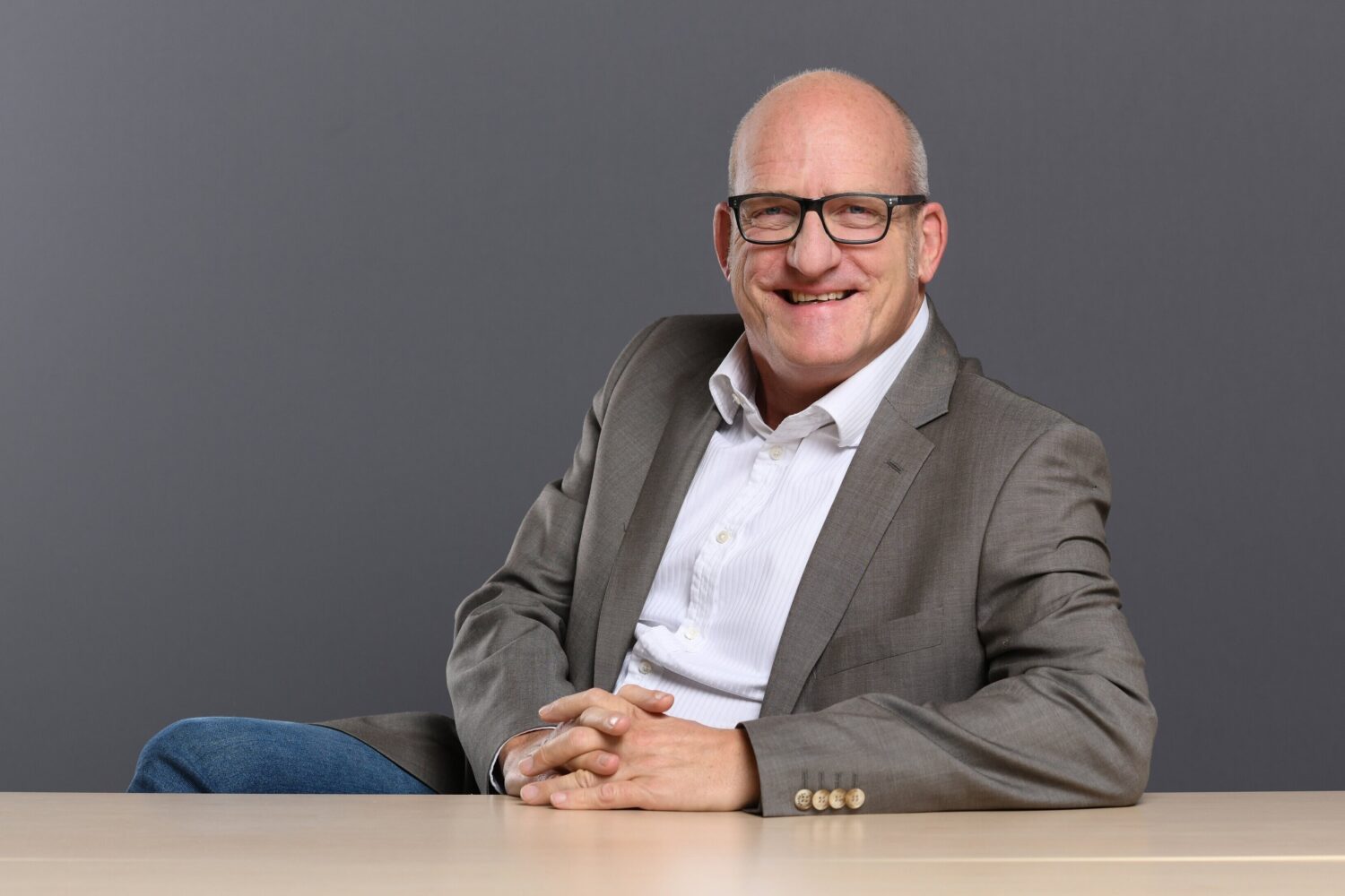 Lächelnder Mann in Anzug sitzt an einem Tisch vor grauem Hintergrund.