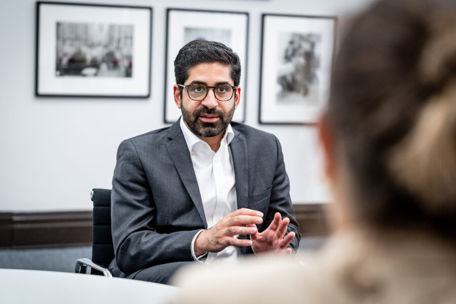 Zwei Personen im Gespräch, eine im Anzug, unscharfe Fotos im Hintergrund.