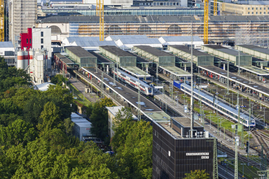 Bahnhof mit mehreren Gleisen, Zügen und Menschen, umgeben von Gebäuden und Bäumen.