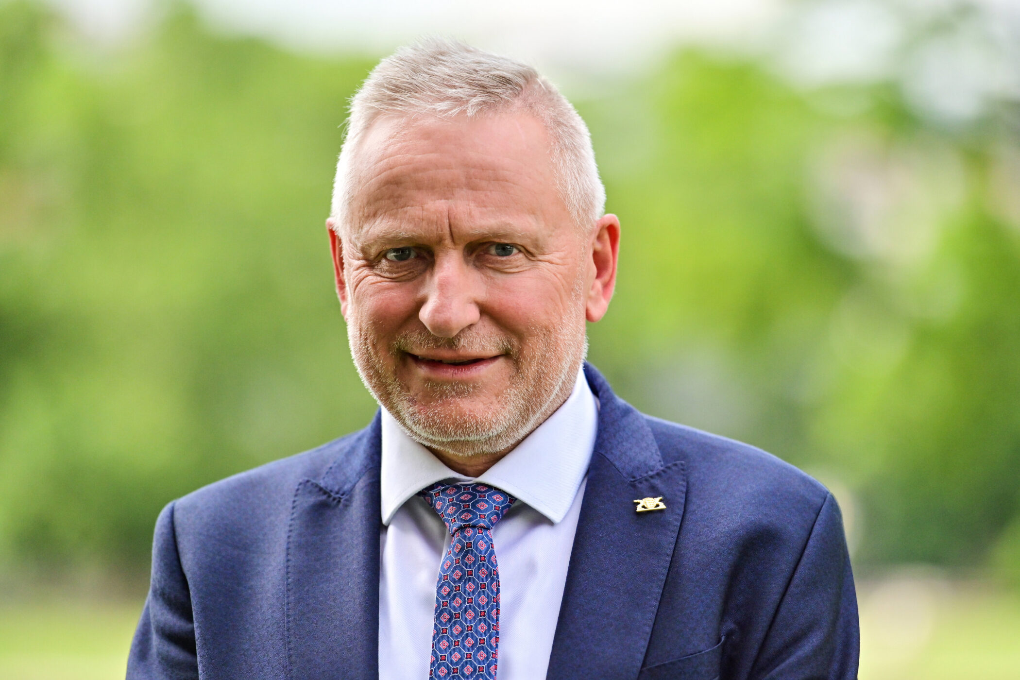 Älterer Mann in Anzug mit Krawatte, lächelnd, unscharfer grüner Hintergrund.