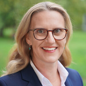 Eine lächelnde Person mit Brille im Freien.