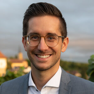 Mann mit Brille und Anzug vor unscharfem Hintergrund.