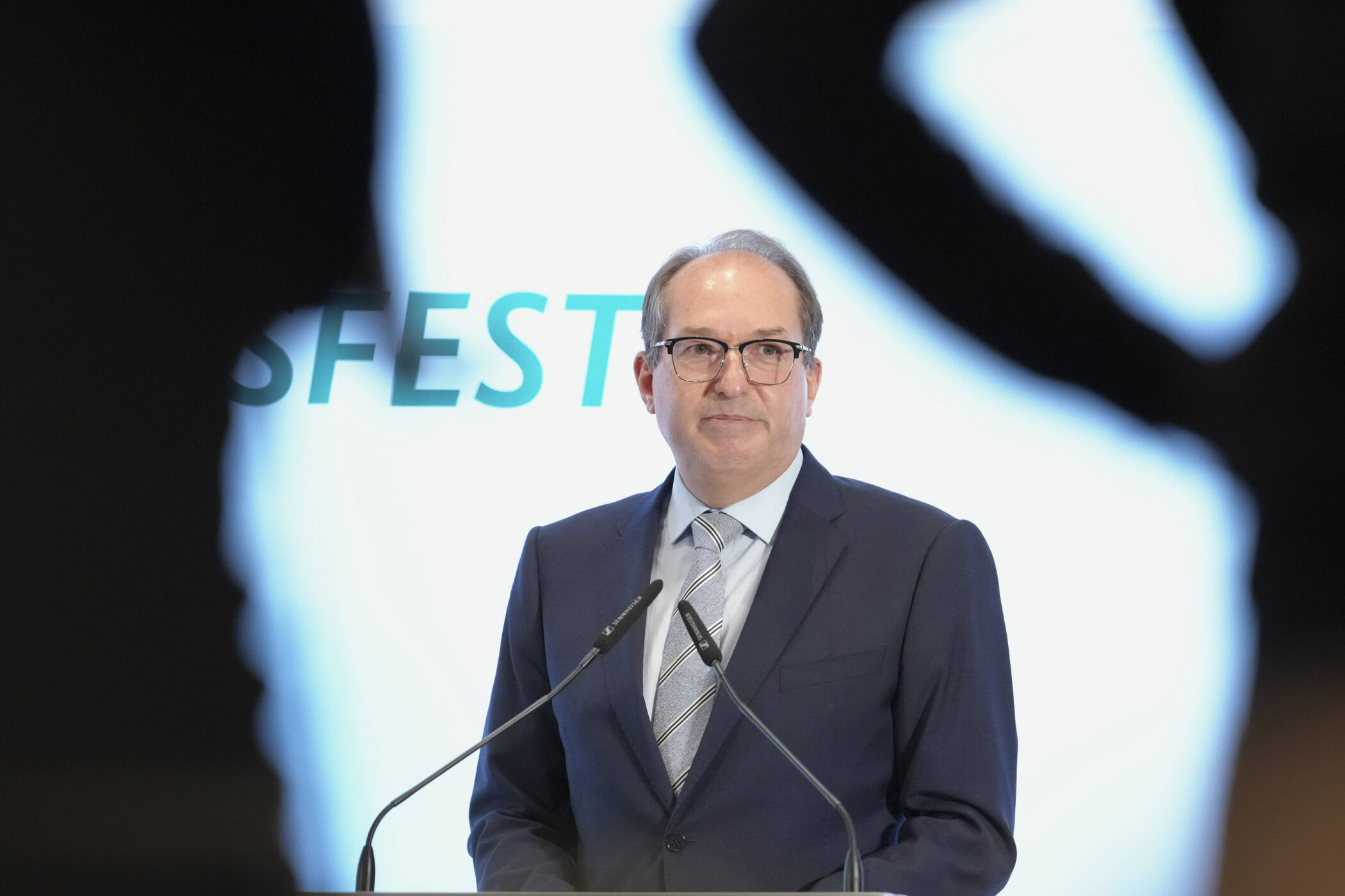 Mann im Anzug spricht vor Mikrofonen, unscharfer Text im Hintergrund.