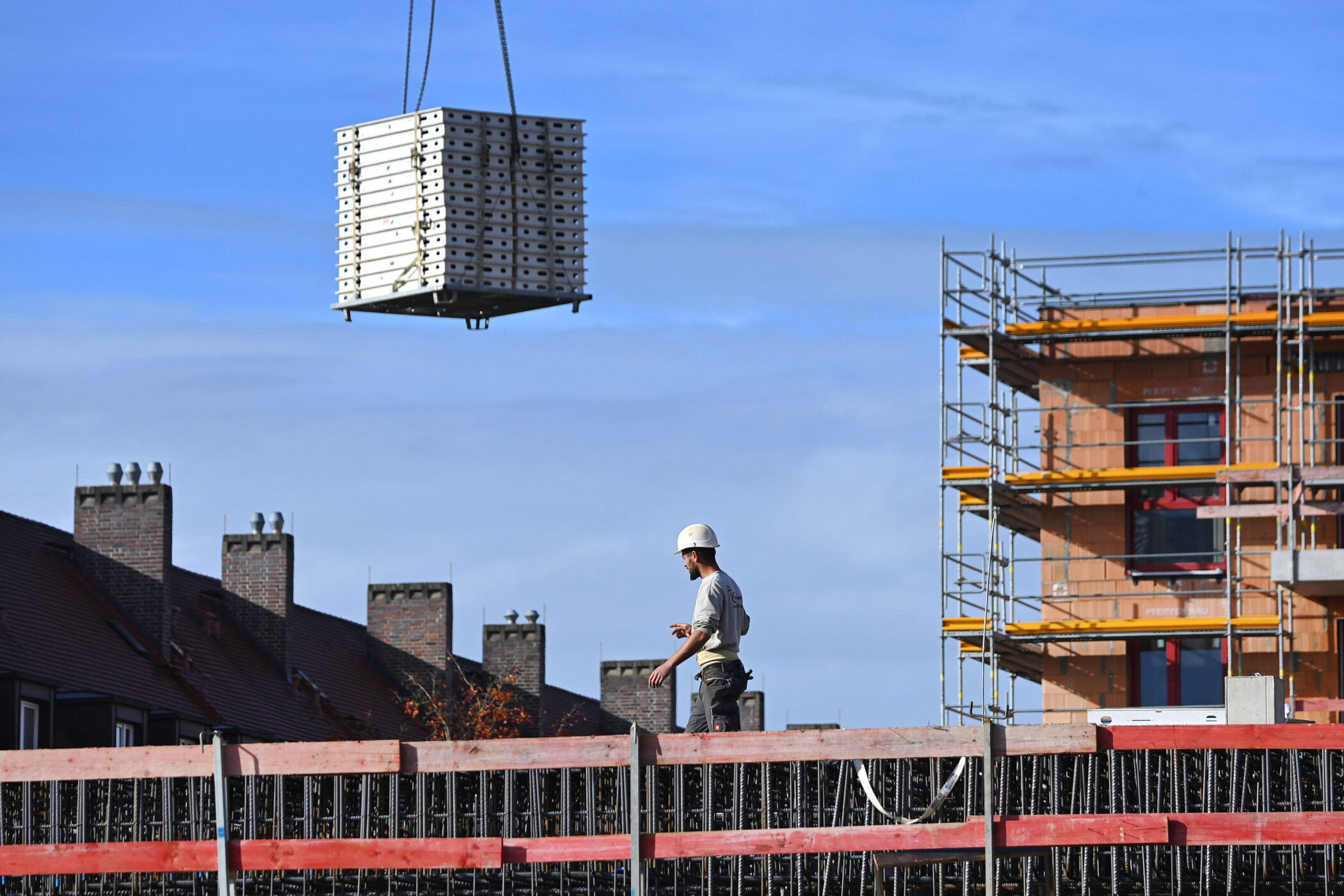 Bauarbeiter mit Helm auf Baustelle, Kran hebt Baumaterial, Gerüst im Hintergrund.