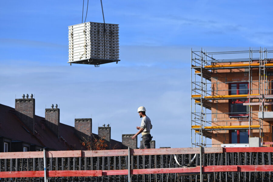 Bauarbeiter mit Helm auf Baustelle, Kran hebt Baumaterial, Gerüst im Hintergrund.