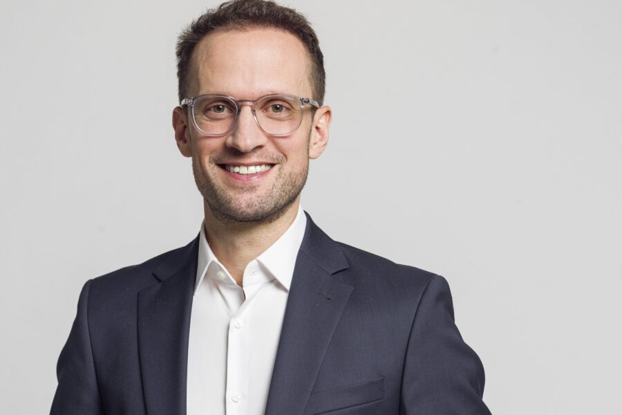 Ein lächelnder Mann mit Brille im Anzug vor grauem Hintergrund.