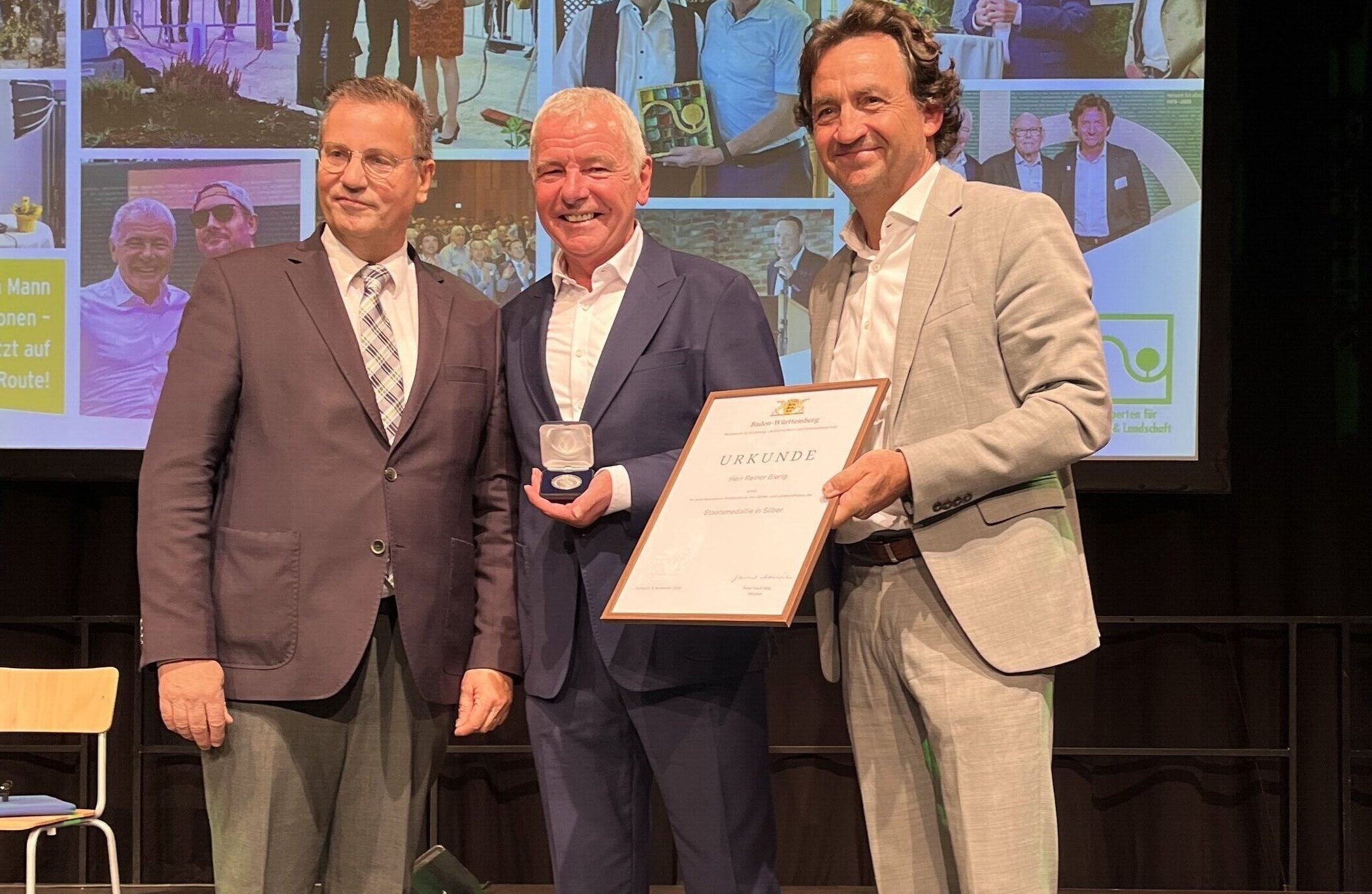 Drei Männer in Anzügen, einer hält eine Urkunde, ein anderer eine Medaille.