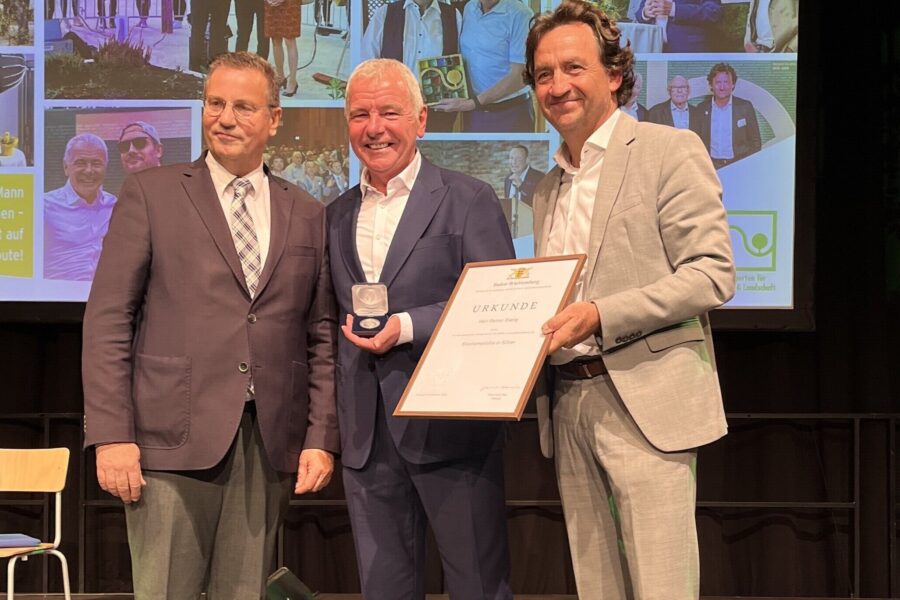 Drei Männer in Anzügen, einer hält eine Urkunde, ein anderer eine Medaille.