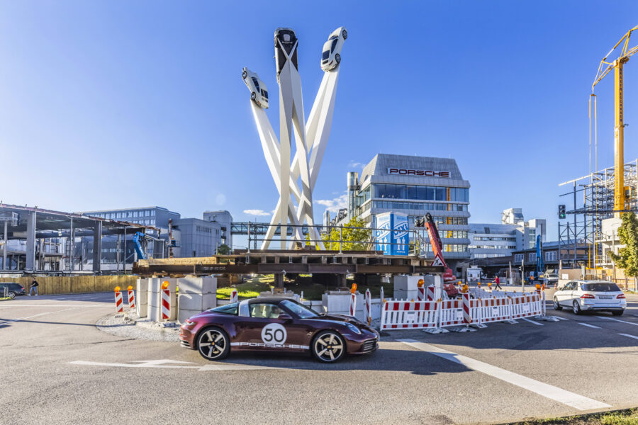 Drei Autos auf hohen Säulen, Gebäude mit Porsche-Logo, vorbeifahrende Autos.