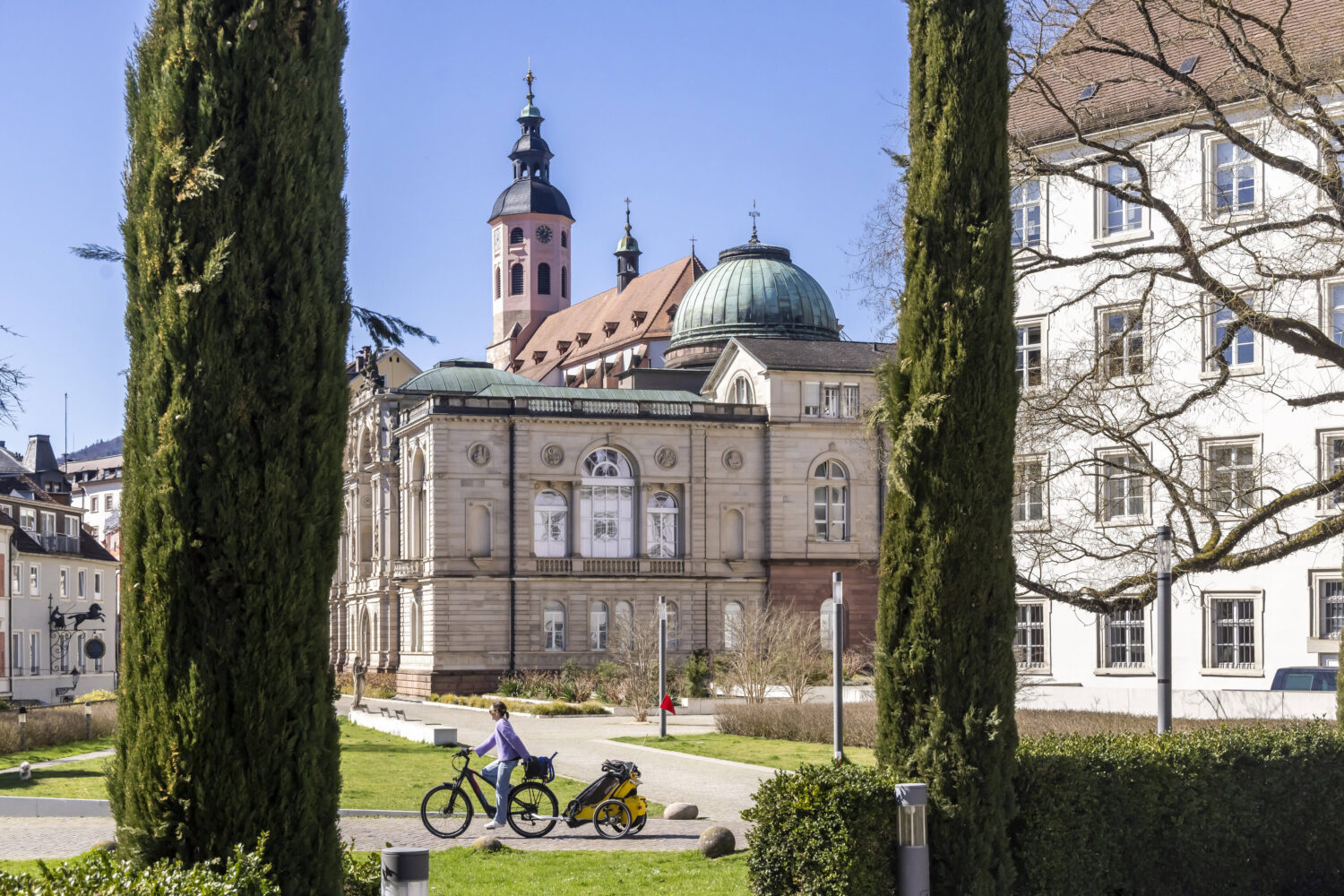 Zwei Zypressen, Gebäude im Hintergrund, Person mit Fahrrad und Anhänger im Vordergrund.