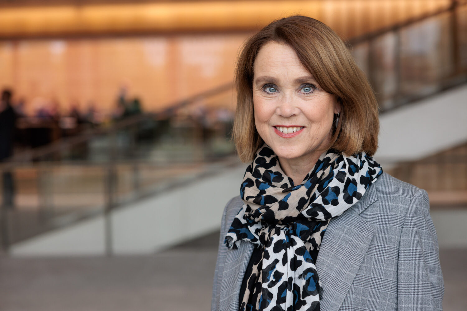 Frau mit schulterlangem Haar und Schal lächelt in die Kamera, unscharfer Hintergrund.