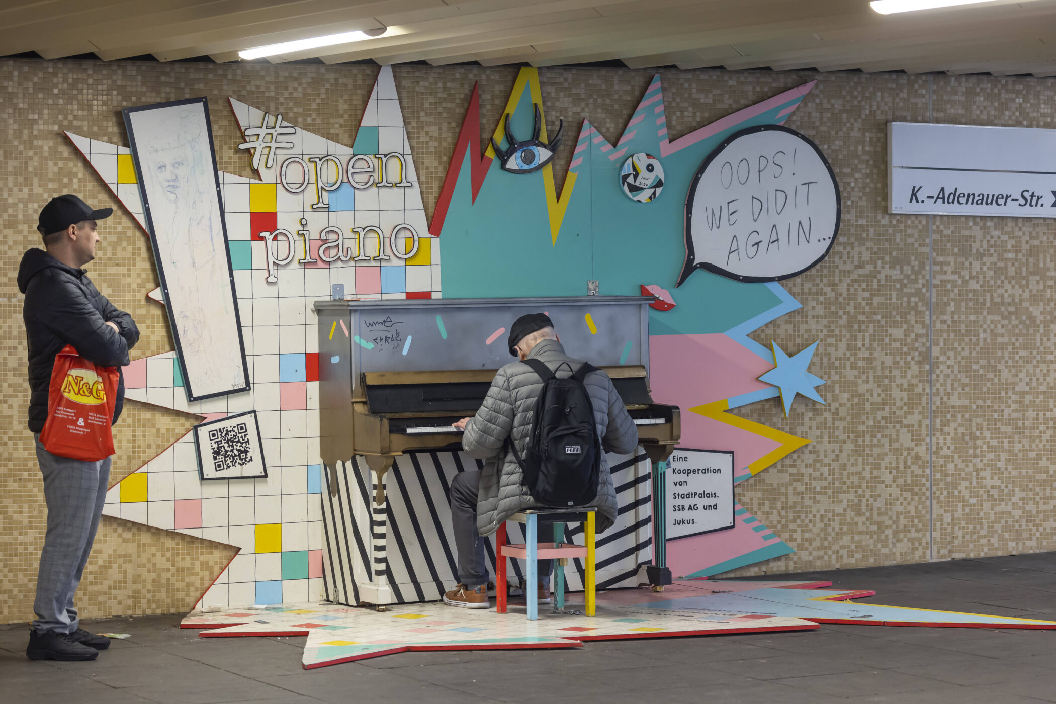 Zwei Männer in einer U-Bahn-Station, einer spielt Klavier, bunte Wandgestaltung.