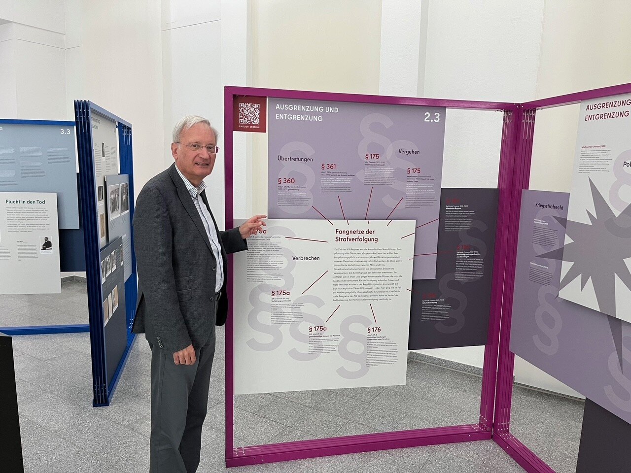 Älterer Mann zeigt auf eine Ausstellungstafel mit Texten und Paragraphen.