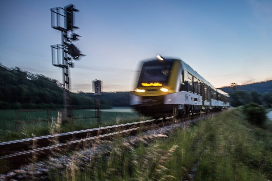 Zug fährt bei Dämmerung auf Gleisen, Signal und Bäume im Hintergrund.