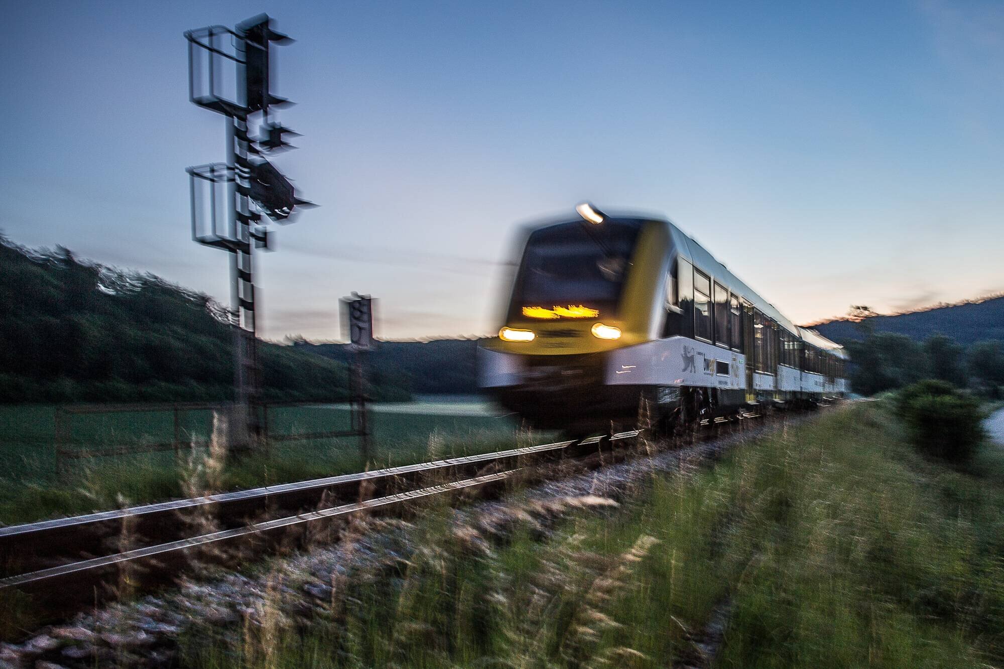 Zug fährt bei Dämmerung auf Gleisen, Signal und Bäume im Hintergrund.