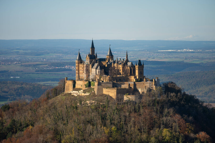 Große Burg auf einem bewaldeten Hügel, umgeben von Landschaft und blauem Himmel.
