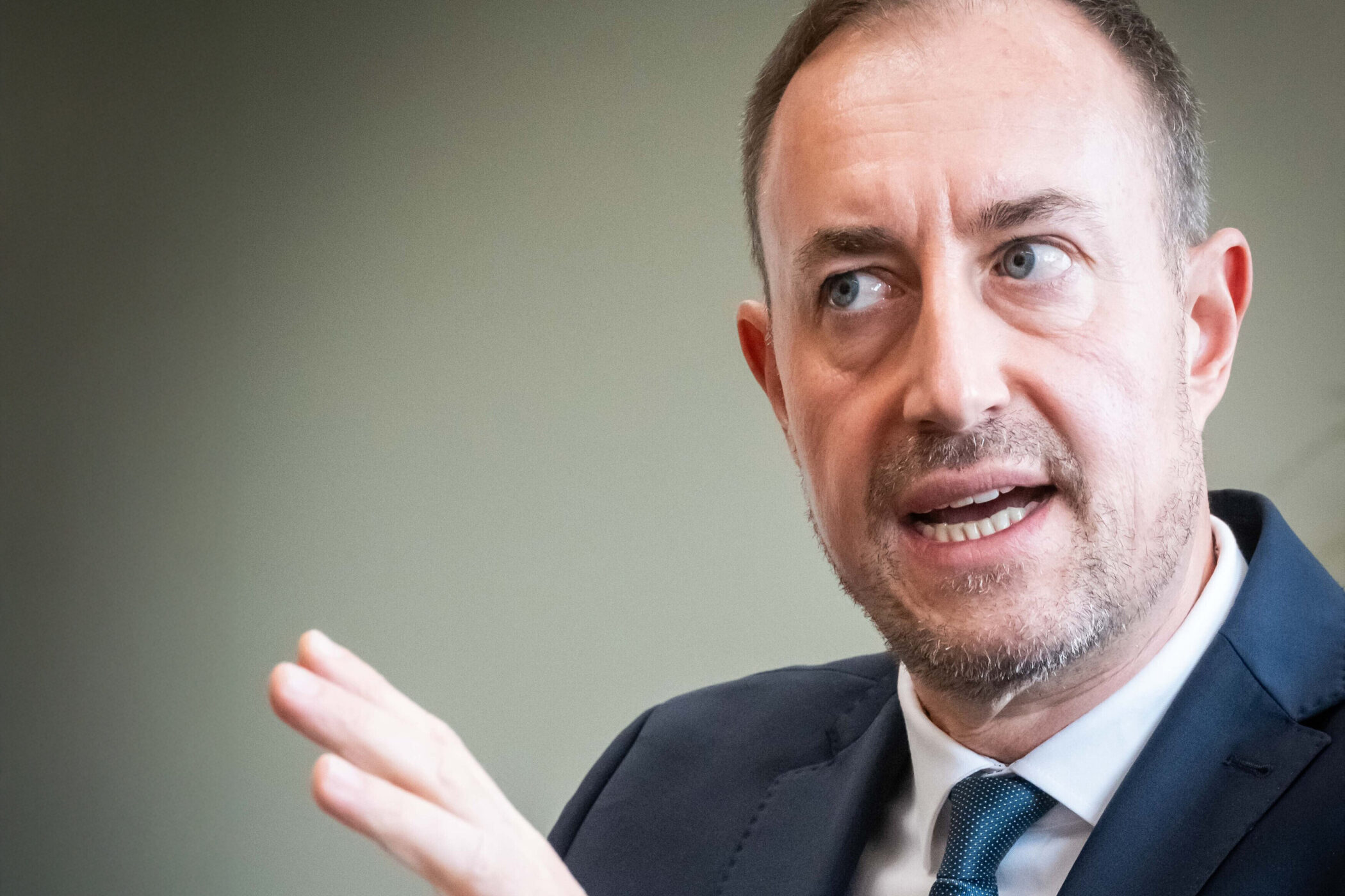 Mann in Anzug spricht, hebt eine Hand, neutraler Hintergrund.