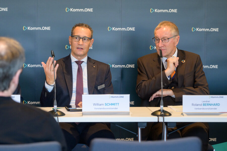 Zwei Männer sitzen an einem Tisch mit Mikrofonen, Hintergrund mit "Komm.ONE"-Logo.