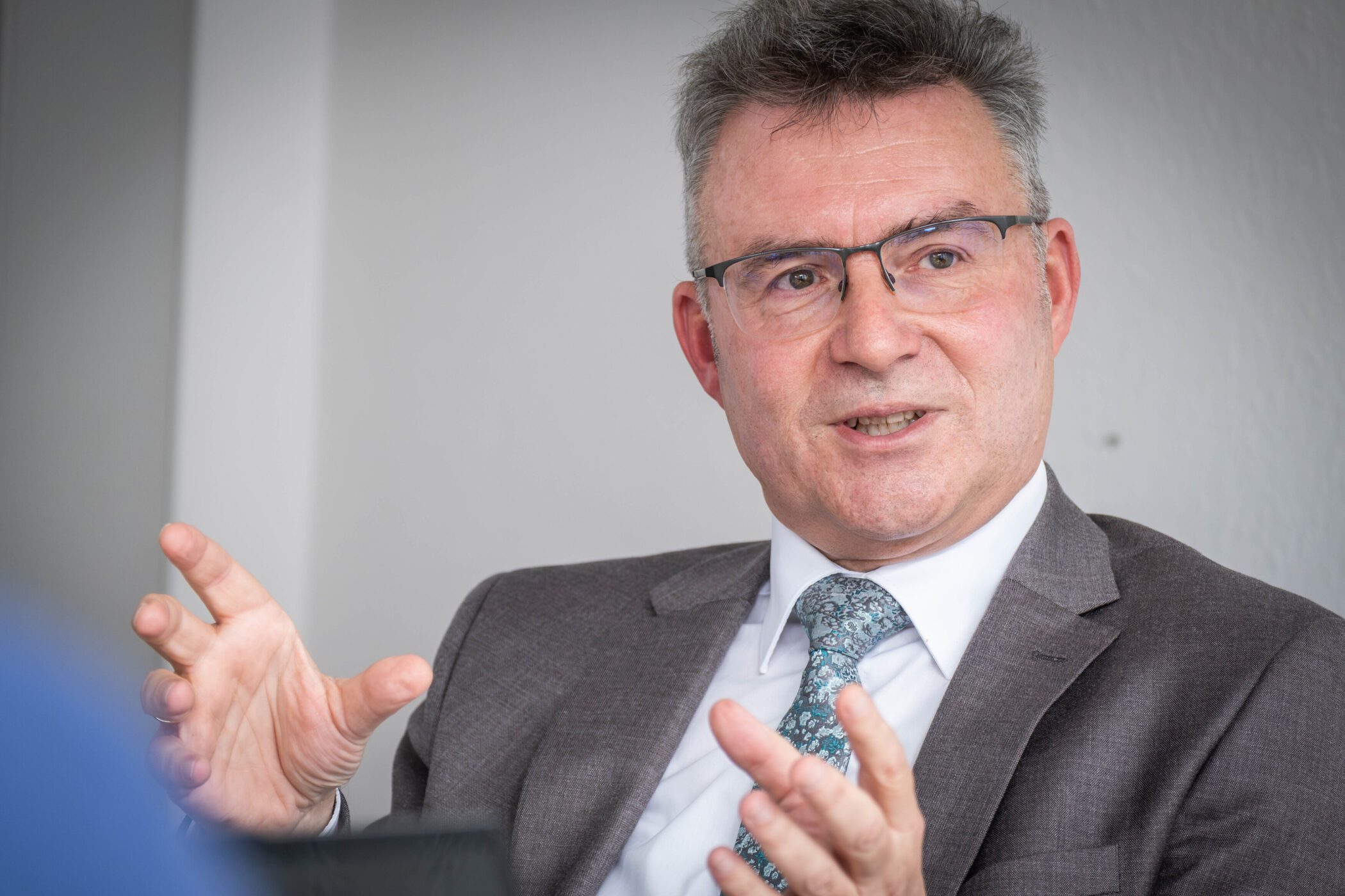 Mann im Anzug spricht gestikulierend, grauer Hintergrund.