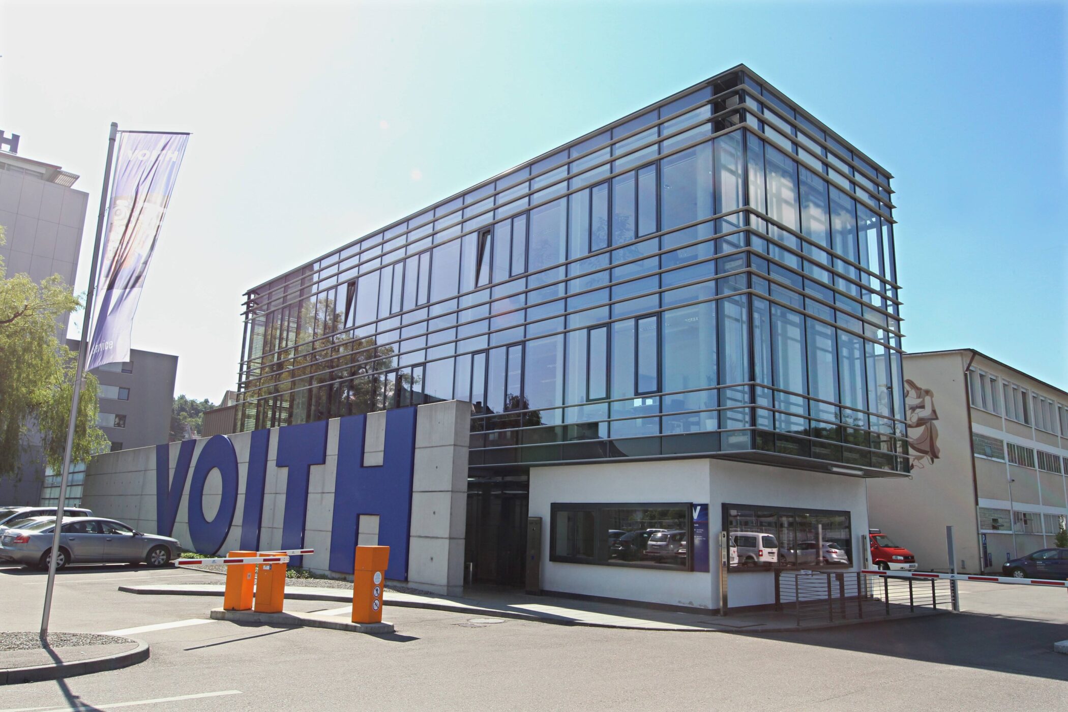 Modernes Gebäude mit Glasfassade, großer Schriftzug "VOITH", Parkplatz im Vordergrund.