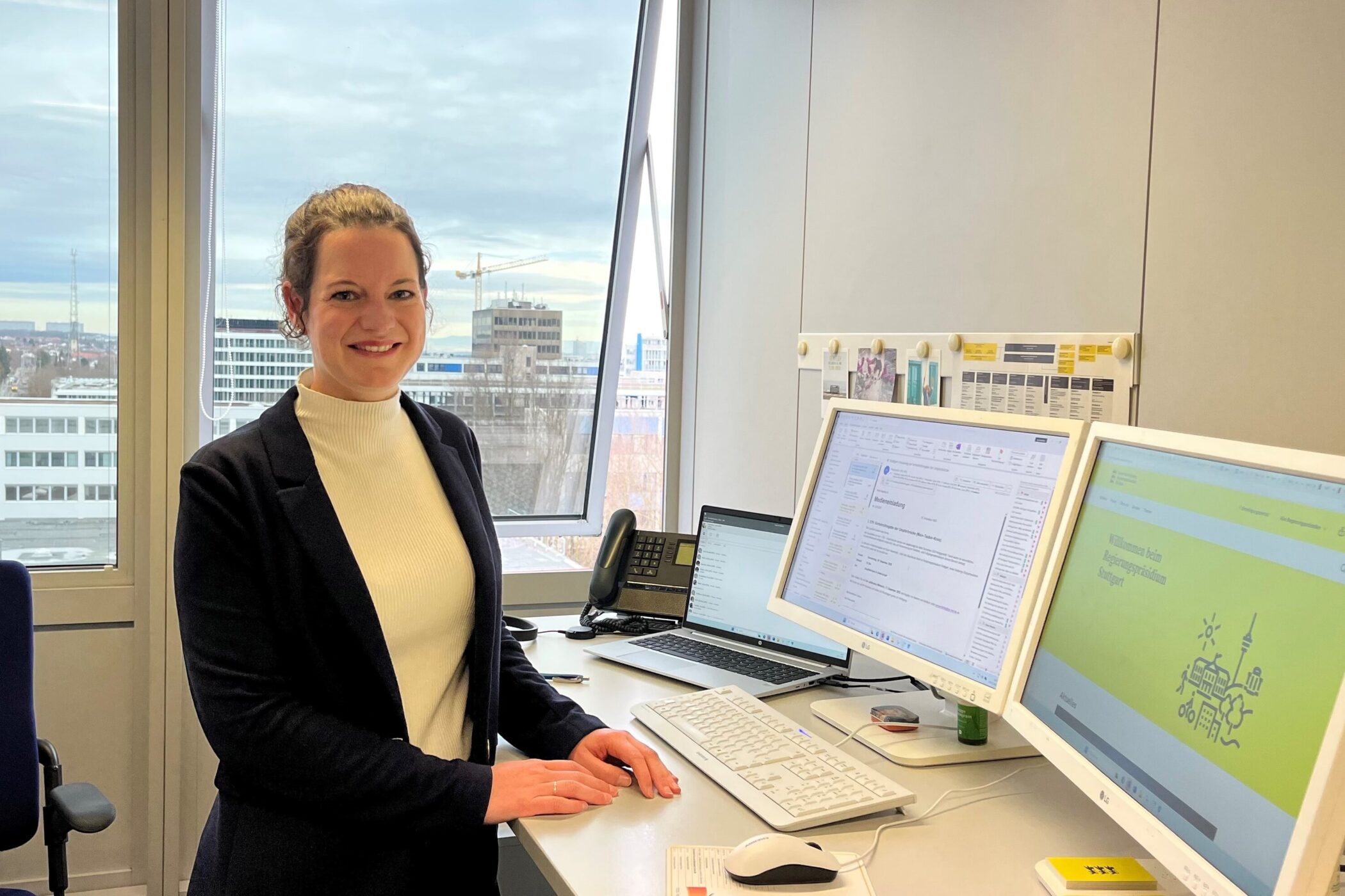 Frau steht lächelnd an einem Schreibtisch mit zwei Monitoren und einem Fenster im Hintergrund.